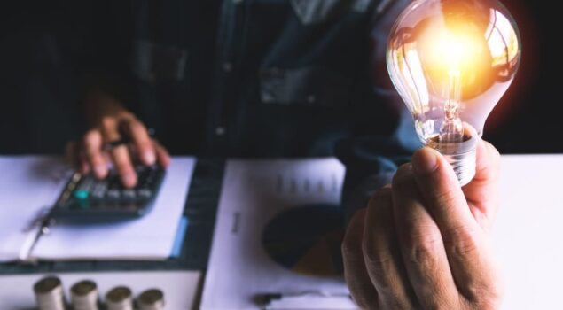 Pessoa segura uma lâmpada acesa enquanto faz cálculos financeiros, representando o impacto da bandeira vermelha na conta de energia.