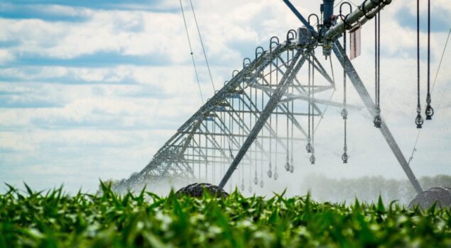 Pivô central de irrigação em funcionamento irrigando uma plantação verde sob céu azul com nuvens.