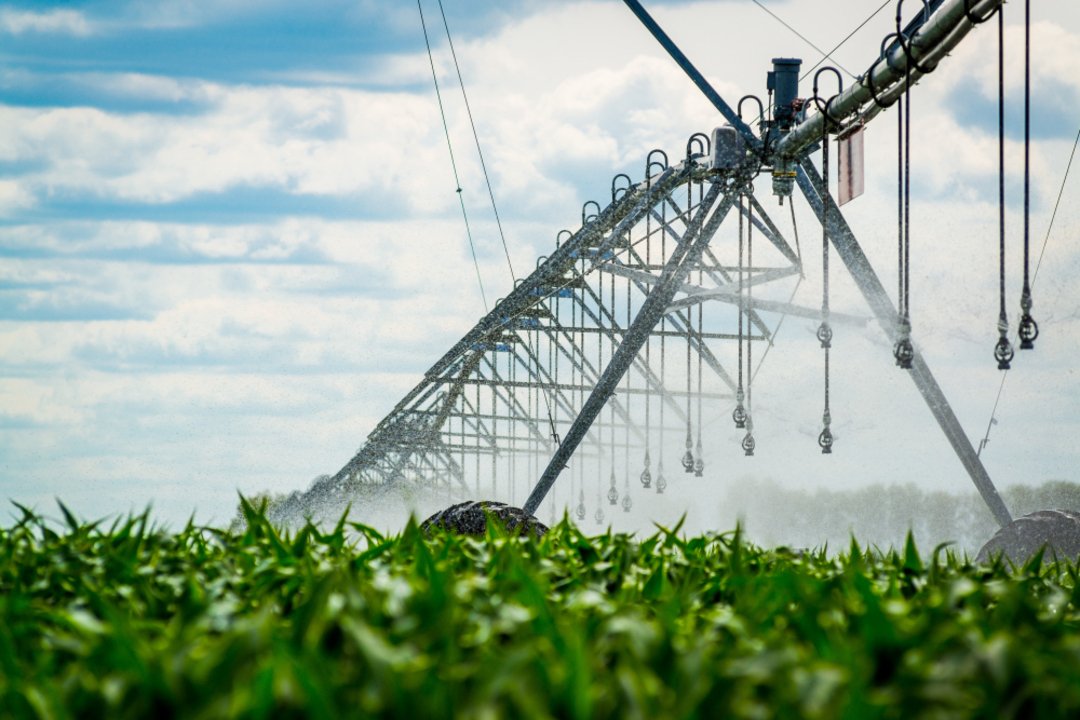 Pivô central de irrigação em funcionamento irrigando uma plantação verde sob céu azul com nuvens.