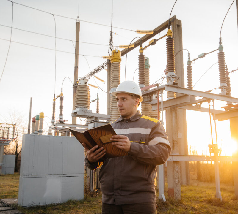 Técnico de engenharia elétrica inspecionando equipamentos de subestação industrial para prevenir falhas energéticas e linha de produção parada.