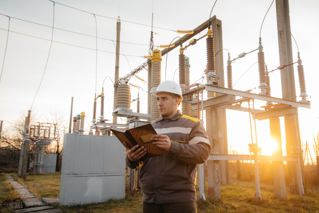 Técnico de engenharia elétrica inspecionando equipamentos de subestação industrial para prevenir falhas energéticas e linha de produção parada.