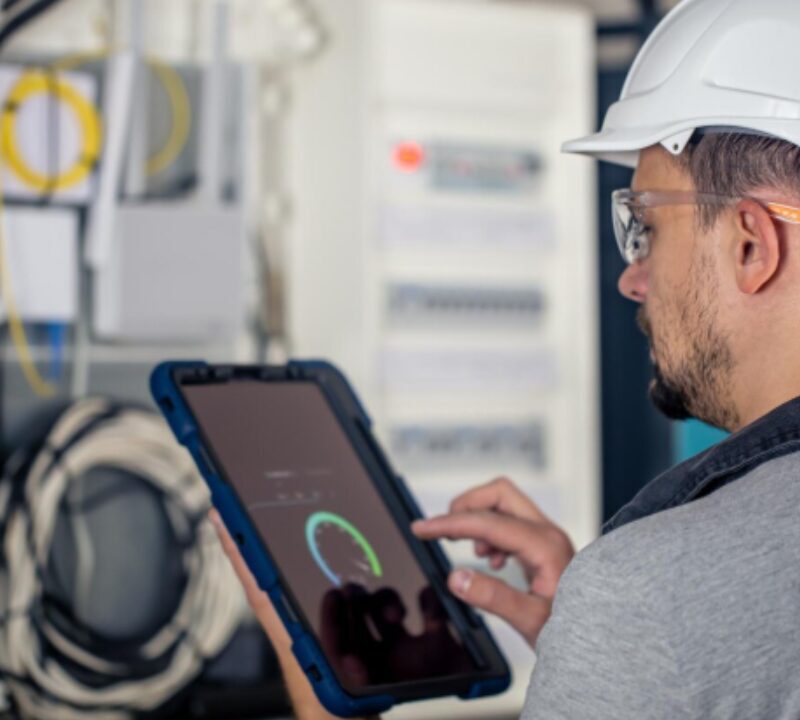 Técnico utilizando tablet para monitoramento e inspeção de sistema elétrico em ambiente industrial.