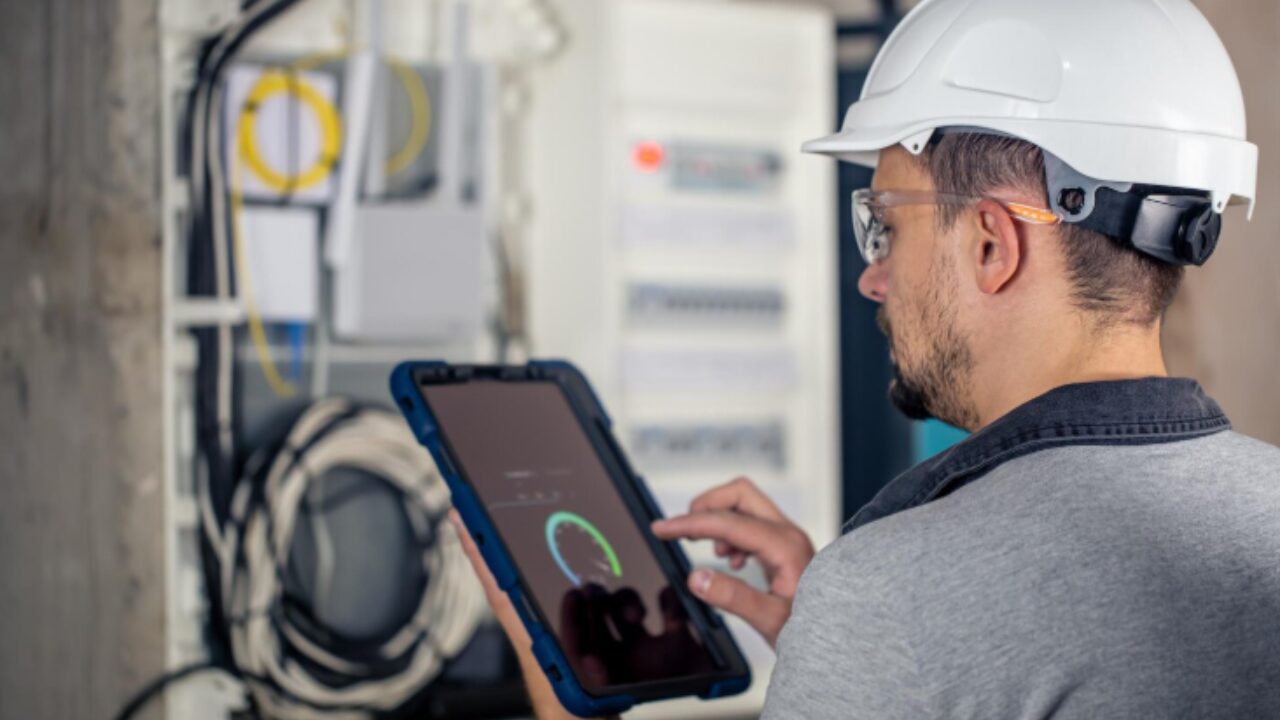 Técnico utilizando tablet para monitoramento e inspeção de sistema elétrico em ambiente industrial.