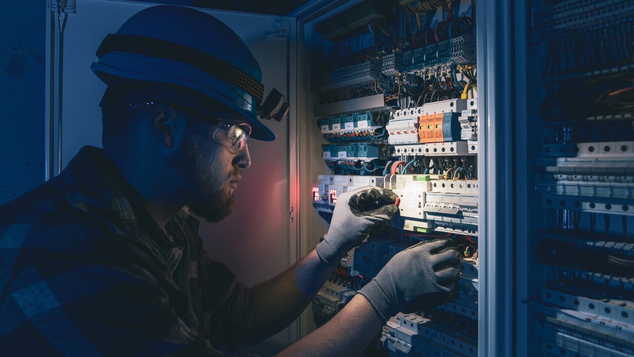 Técnico realiza manutenção em painel elétrico utilizando equipamentos de proteção individual e ferramentas de medição.