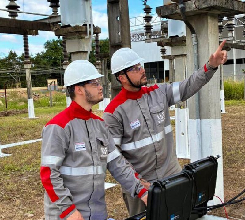 Dois técnicos com equipamentos de proteção realizam inspeção em uma subestação elétrica, analisando componentes da instalação.