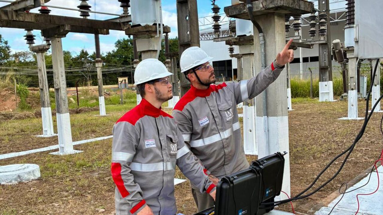 Dois técnicos com equipamentos de proteção realizam inspeção em uma subestação elétrica, analisando componentes da instalação.